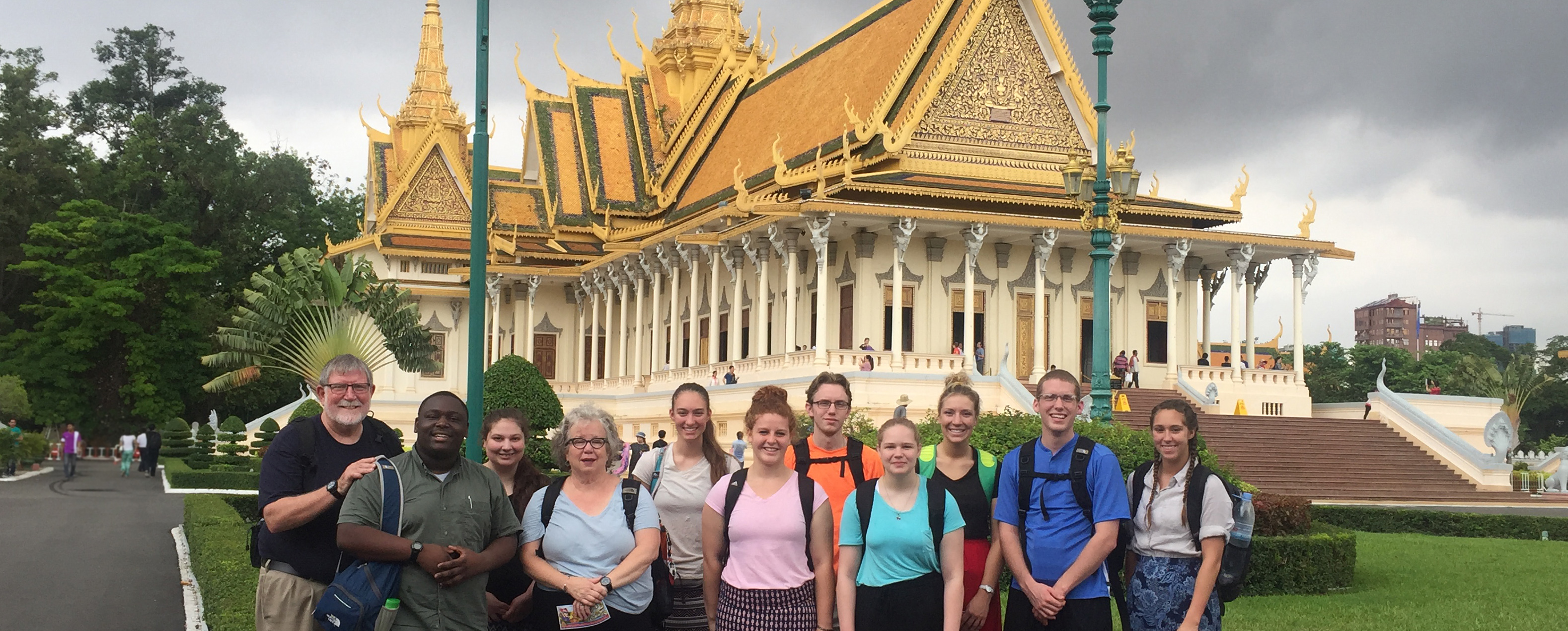 11 individuals from Defiance College wearing all different and brightly colored shirts with backpacks. They are gathered together to smile at the camera, standing in front of a large pale yellow building with a tall golden roof. The building is the Silver Pagoda in Cambodia.