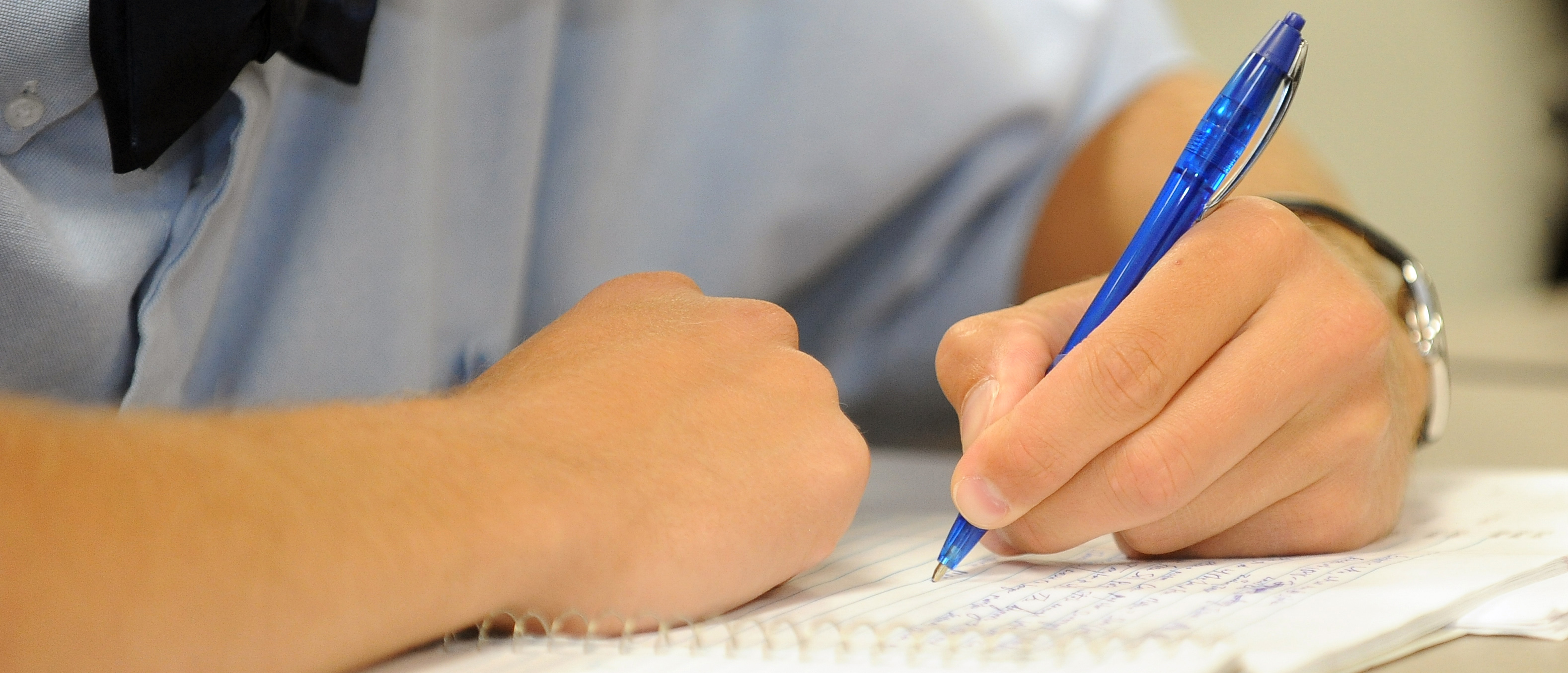Student's arms leaning on desk and writing in a spiral notebook with a blue pen