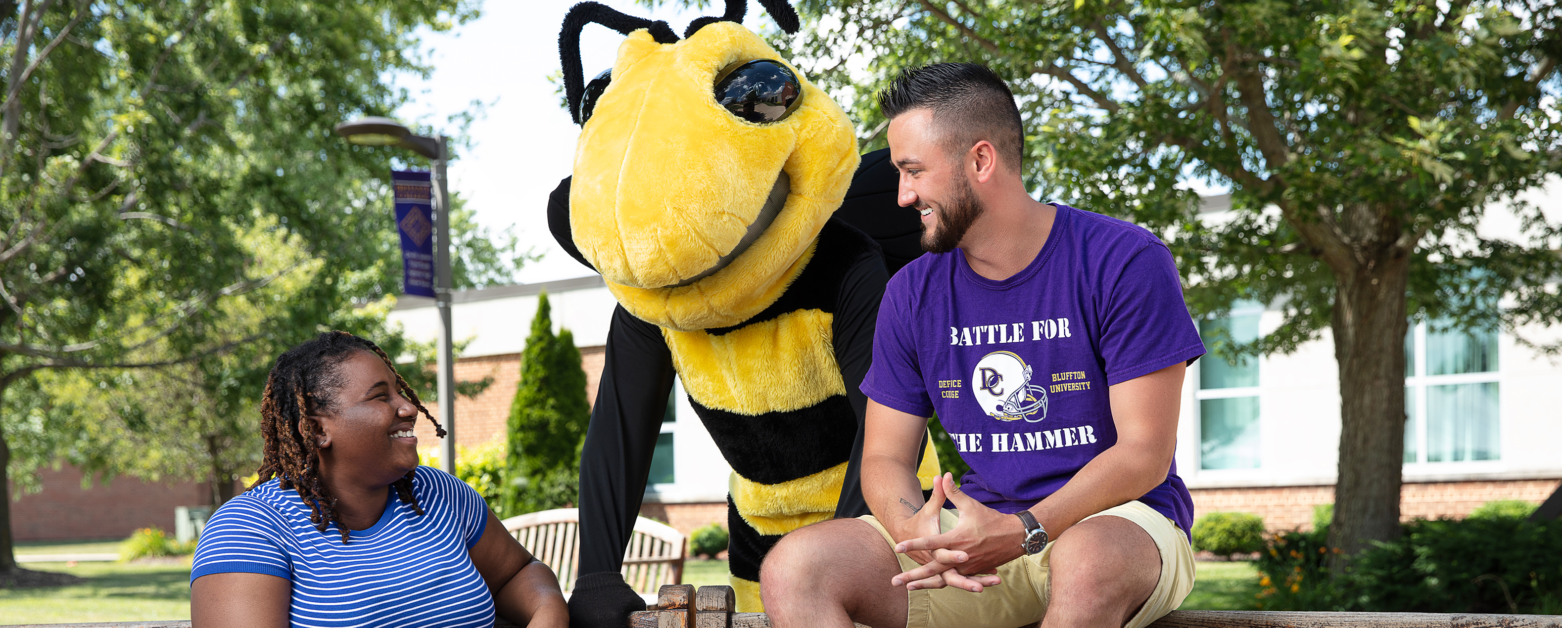 Two students on a wooden bench by the campus sidewalk with the campus yellow jacket mascot