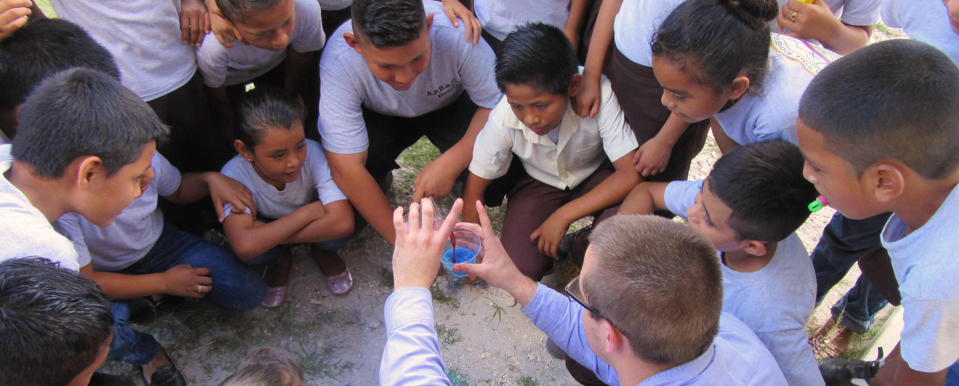 14 kids of varying ages gathered in a circle around a young man who is holding a cup filled with blue liquid.